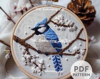 May include: Embroidered hoop art featuring a blue jay perched on a branch with white blossoms. The embroidery is done in shades of blue, white, and brown on a white fabric.