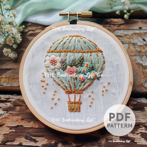 May include: Embroidered hot air balloon with floral accents in a wooden embroidery hoop. The balloon is light blue with gold accents and the flowers are pink, white, and blue. The text "THE Embroidery Loft" is visible on the hoop.
