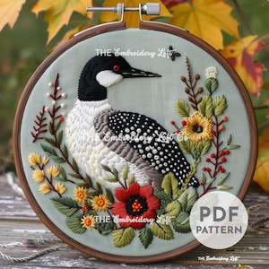 May include: Embroidered hoop art featuring a loon bird with a black head and white and gray body, surrounded by colorful flowers and greenery. The hoop is made of wood and has a light blue background. The text "THE Embroidery Loft" is visible on the hoop.