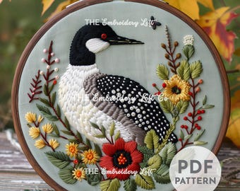 Könnte beinhalten: Stickbild mit Stickerei, das einen  Seetaucher mit schwarzem Kopf und weißem und grauem Körper zeigt, umgeben von bunten Blumen und Grün. Der Stickrahmen ist aus Holz und hat einen hellblauen Hintergrund. Der Text "THE Embroidery Loft" ist auf dem Rahmen sichtbar.