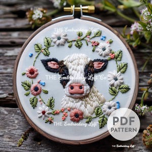 May include: Embroidered hoop art featuring a white and brown cow with black eyes and a pink nose. The cow is surrounded by a wreath of pink, white, and green flowers. The text "THE Embroidery Loft" appears three times around the hoop.