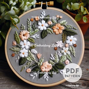 Puede incluir: Un aro de bordado gris con un diseño floral bordado en hilo blanco, melocotón y verde. El diseño presenta una corona de flores y hojas. El texto "THE Embroidery Loft" aparece tres veces en el aro. El texto "PDF PATTERN" aparece en la esquina inferior derecha del aro.
