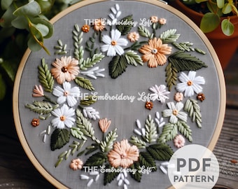 May include: A gray embroidery hoop with a floral design embroidered in white, peach, and green thread. The design features a wreath of flowers and leaves. The text "THE Embroidery Loft" appears three times on the hoop. The text "PDF PATTERN" appears in the lower right corner of the hoop.