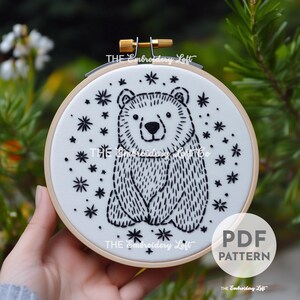 May include: A black and white embroidery pattern of a bear sitting with stars around it. The pattern is on a white hoop with the text "THE Embroidery Loft" printed on the bottom.