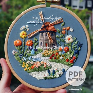 May include: Embroidered hoop art depicting a windmill with a red roof and a path leading to it. The windmill is surrounded by a variety of colourful flowers and greenery. The text "THE Embroidery Loft Co" is visible in the image.