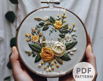 May include: A close-up of a hoop with a floral embroidery design. The embroidery features yellow and white roses, green leaves, and small white flowers. The hoop is held in a person's hands. The text "THE Embroidery Loft" and "PDF PATTERN" is visible in the image.