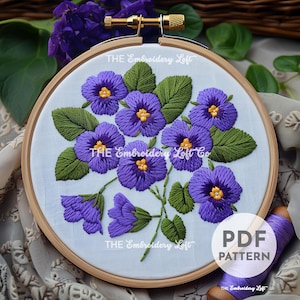 May include: A PDF pattern for embroidery featuring a bouquet of purple pansies with green leaves. The pattern is displayed in a wooden embroidery hoop.