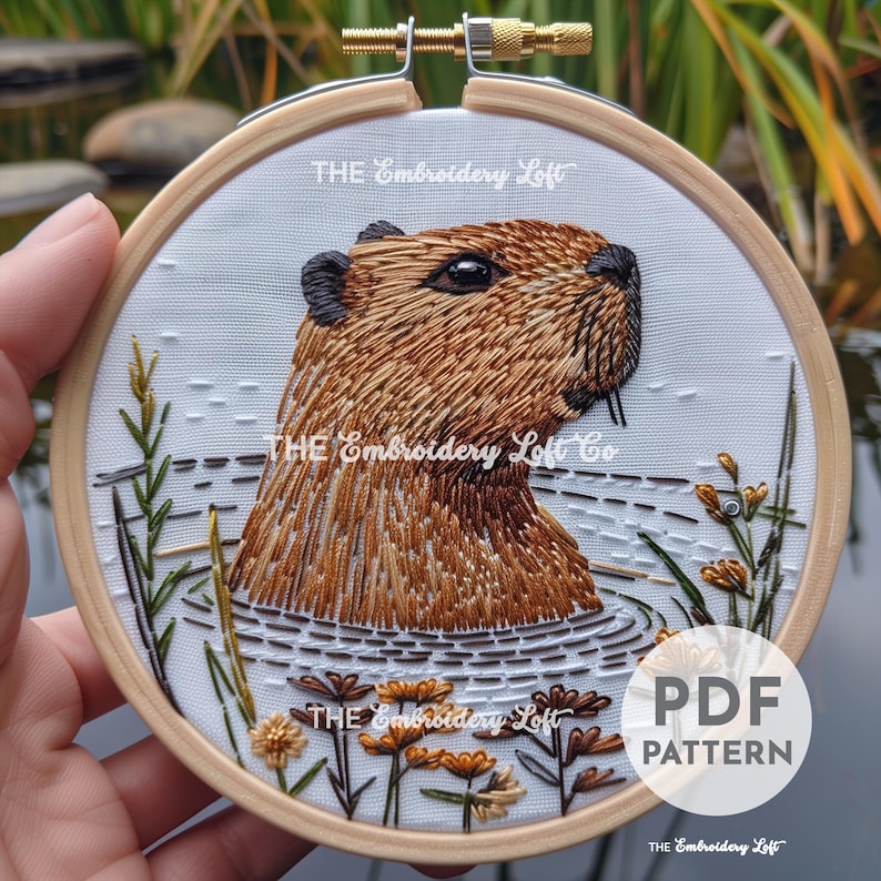 K&ouml;nnte beinhalten: Stickmuster eines Capybaras in einem Teich mit gr&uuml;nen Schilf und gelben Blumen. Das Muster befindet sich in einem h&ouml;lzernen Stickrahmen. Der Text "THE Embroidery Loft Co" ist auf dem Muster sichtbar.