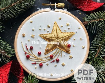 May include: A gold star with a red and gold tail, embroidered on white fabric in a wooden embroidery hoop. The star is surrounded by small gold and white beads. The text "THE Embroidery Loft Co." is visible on the fabric.
