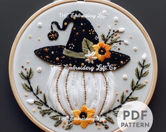 May include: Embroidered hoop art featuring a white pumpkin with a black witch hat adorned with yellow and white flowers and green leaves. The text "THE Embroidery Loft Co" is stitched on the pumpkin.