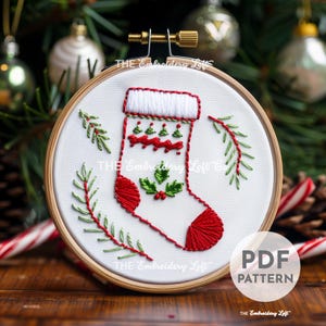 May include: A white embroidery hoop with a red and green embroidered Christmas stocking with holly and a sprig of greenery. The text "THE Embroidery Loft Co." is embroidered on the hoop.