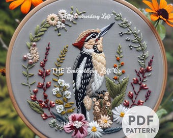 May include: Embroidered hoop art featuring a detailed depiction of a woodpecker perched on a branch surrounded by delicate floral embroidery. The hoop is made of wood and has a gray fabric background. The text "THE Embroidery Loft Co." is visible in the image.