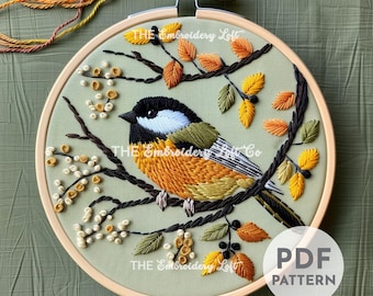 May include: Embroidery pattern featuring a small bird with yellow, orange, and green plumage perched on a branch with white berries and brown leaves. The pattern is in a hoop with a light green background. The text "THE Embroidery Loft" appears three times on the hoop.