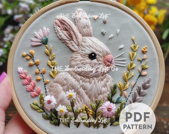 Puede incluir: Arte de bordado en aro que presenta un conejo blanco sentado en un campo de flores rosas, blancas y amarillas. El aro es de madera y tiene un fondo de tela gris claro. El texto "THE Embroidery Loft" es visible en la parte inferior del aro.