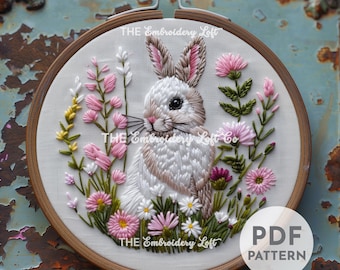 Puede incluir: Arte de bordado en aro con un conejo blanco con orejas rosas sentado en un campo de flores rosas y blancas. El aro es de madera y tiene un fondo de tela blanca. El texto "THE Embroidery Loft" está impreso en el aro.