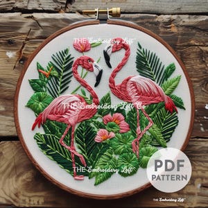 May include: Embroidery pattern featuring two pink flamingos standing in a lush green tropical foliage with pink flowers. The pattern is in a hoop and says "PDF PATTERN" at the bottom.