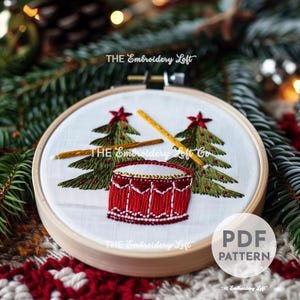 May include: Embroidered hoop art featuring a red drum with two yellow drumsticks and two green Christmas trees with yellow stars on top. The hoop is made of wood and the fabric is white. The text "THE Embroidery Loft" is visible on the hoop.