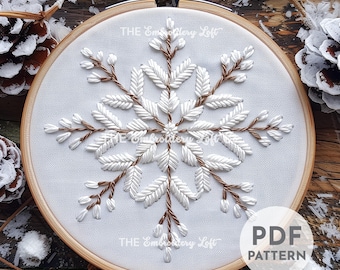 May include: White snowflake embroidery pattern on a white fabric in a wooden embroidery hoop. The pattern is made with white satin stitch and brown backstitch. The text "PDF PATTERN" is in the lower right corner of the image.