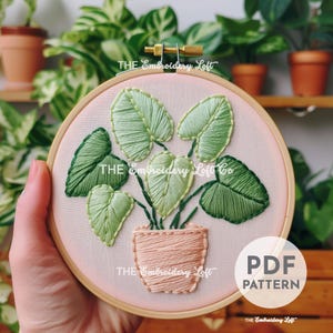 Puede incluir: Patrón de bordado para una planta en maceta con hojas verdes. El patrón está en un aro de bordado rosa e incluye el texto "THE Embroidery Loft Co."