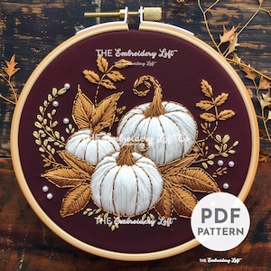 Peut inclure: Cerceau de broderie avec un fond en tissu bordeaux représentant trois citrouilles blanches avec des détails brodés en or. Les citrouilles sont entourées de feuilles et de vignes brodées en or. Le texte "THE Embroidery Loft" apparaît deux fois sur le cerceau.