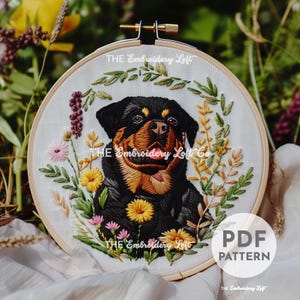 Puede incluir: Diseño bordado de un perro Rottweiler rodeado de flores verdes, amarillas y rosas en un aro de bordado de madera. El texto "THE Embroidery Loft Co" es visible en el diseño.