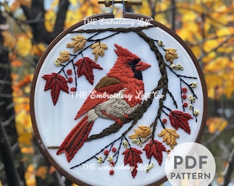 May include: Embroidered design of a cardinal bird perched on a branch with red, yellow, and brown leaves. The embroidery is in a white hoop with a brown wooden frame. The text "THE Embroidery Loft Co" is visible in the image.