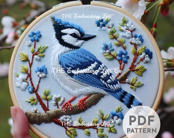 May include: Hand-stitched embroidery art featuring a blue jay perched on a branch with blue and white flowers. The embroidery is set in a wooden hoop. The text "THE Embroidery Loft" and "PDF PATTERN" are visible.