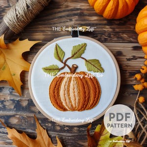 Peut inclure: Citrouille brodée avec des feuilles vertes sur un fond blanc dans un cercle de broderie en bois. Le cercle est sur une surface en bois avec des feuilles d'automne et une citrouille.