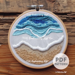 May include: Embroidered hoop art featuring a blue and white wave design with a sandy beach at the bottom. The hoop is made of wood and has a white fabric background. The text "THE Embroidery Loft Co" is visible in the centre of the hoop.