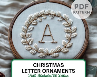 Pode incluir: Enfeite de letra de Natal bordado em um bastidor de madeira. A letra "A" é bordada com fio marrom, rodeada por uma coroa de folhas brancas e marrons. O texto "CHRISTMAS LETTER ORNAMENTS Full Alphabet 26 Letters" é exibido em um retângulo verde.
