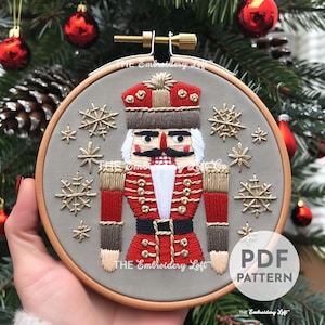 May include: Embroidered hoop art featuring a Nutcracker soldier in red, gold, and white with a gray background and gold snowflakes. The text "THE Embroidery Loft" is at the bottom of the hoop.