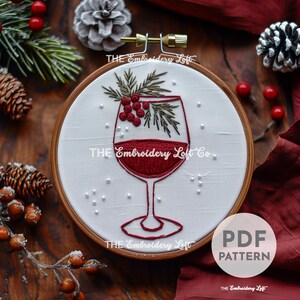 Puede incluir: Diseño bordado de una copa de vino tinto con una rama de vegetación y bayas rojas. El diseño está sobre un fondo de tela blanca en un aro de bordado de madera. El texto "THE Embroidery Loft Co." es visible en la imagen.