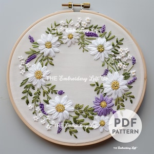 May include: A white embroidery hoop with a floral design of white daisies and purple lavender. The design is stitched with white and purple embroidery floss on a white fabric. The hoop is made of wood and has a natural finish. The text "PDF PATTERN" is printed in black on a white background in the lower right corner of the image.