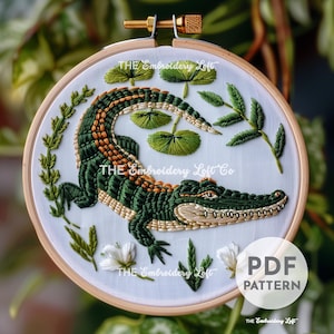 Puede incluir: Patrón de bordado para un cocodrilo verde con flores blancas y hojas verdes. El patrón está en un aro y dice "THE Embroidery Loft Co" y "PDF PATTERN".