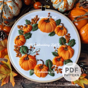 May include: Embroidered hoop art featuring a wreath of orange pumpkins and green leaves. The hoop is made of wood and has a white fabric background. The text "THE Embroidery Loft Co." is visible on the fabric.