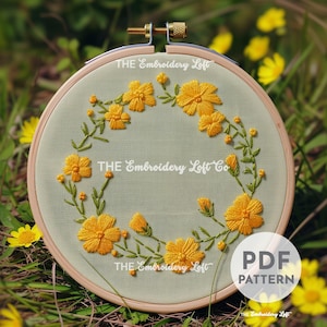 May include: A green embroidery hoop with a floral wreath design featuring yellow flowers and green leaves. The text "THE Embroidery Loft Co" is embroidered in white on the fabric. The hoop is sitting on a bed of green grass.