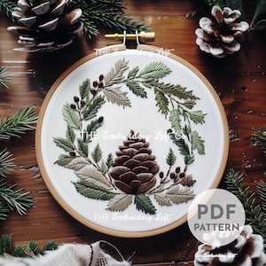 May include: Embroidered hoop art featuring a pine cone surrounded by green and brown foliage. The hoop is made of wood and has a white fabric background. The text "THE Embroidery Loft" is visible at the bottom of the hoop.