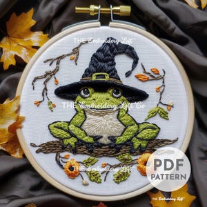 Könnte beinhalten: Stickerei mit einem grünen Frosch, der einen schwarzen Hexenhut mit braunem Rand trägt. Der Frosch sitzt auf einem Ast mit orangefarbenen und braunen Blumen. Das Design ist von einem weißen Hintergrund und einem hölzernen Stickrahmen umgeben.