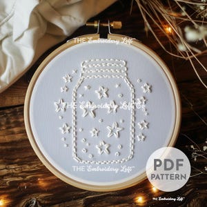 Op de afbeelding: Wit borduurpatroon van een glazen pot gevuld met sterren op een lichtblauwe achtergrond. Het patroon bevindt zich in een houten borduurring. De tekst "THE Embroidery Loft" is op de ring gedrukt en de tekst "PDF PATTERN" is onder de ring gedrukt.