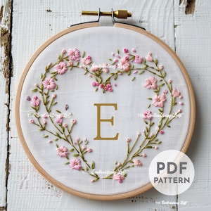 May include: Embroidery hoop with a white fabric background and a pink floral design in the shape of a heart. The letter "E" is embroidered in gold in the center of the heart. The text "PDF PATTERN" is printed in black at the bottom of the hoop.