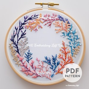 May include: A colourful embroidery hoop featuring a floral wreath design. The wreath is made up of various shades of blue, pink, orange, and gold thread. The text "THE Embroidery Loft Co." is visible in the centre of the hoop.