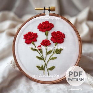Puede incluir: Carnaciones bordadas en rojo y verde sobre tela blanca en un aro. El patrón de bordado está disponible para descargar en PDF. "THE Embroidery Loft" está impreso en la tela.