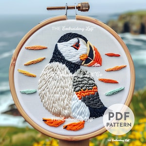 May include: Embroidered pattern of a puffin with orange, white, black, and gray thread on a white fabric in a wooden embroidery hoop. The text "THE Embroidery Loft" is at the top of the hoop and "PDF PATTERN" is at the bottom right of the hoop.