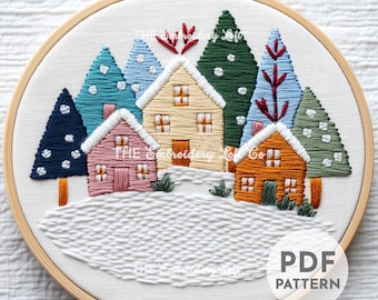 Peut inclure: Scène de village d'hiver brodée avec des maisons, des arbres et de la neige. La broderie est réalisée dans une variété de couleurs, notamment le bleu, le vert, le rouge et le jaune. Le motif est encadré dans un cercle en bois. Le texte "PDF PATTERN" est imprimé dans le coin inférieur droit de l'image.