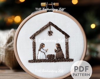 May include: Embroidered hoop art featuring a brown wooden hoop with a white fabric background. The fabric has an embroidered image of a brown manger with a star above it. Two figures, a woman in white and a man in brown, are standing inside the manger. The text "THE Embroidery Loft Co." is embroidered in brown thread below the manger.