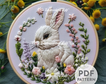Puede incluir: Arte de bordado en aro con un conejo blanco sentado en un campo de flores rosas, moradas y verdes. El aro es de madera y tiene un fondo rosa claro. El texto "THE Embroidery Loft" es visible en la parte superior e inferior del aro.