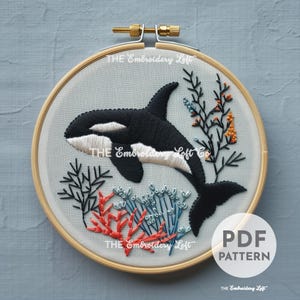 Puede incluir: Patrón de bordado con una orca en blanco y negro nadando a través de corales y algas marinas. El patrón está en un aro de bordado de madera e incluye el texto "PDF PATTERN" y "THE Embroidery Loft".