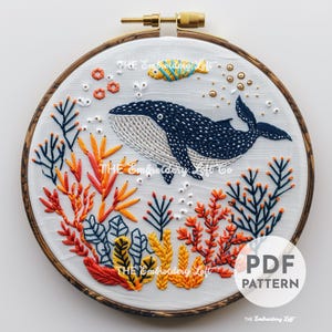以下が含まれることがあります： 刺繍されたフープアートで、サンゴ礁の中で泳ぐクジラが描かれています。クジラは青と白で、黒い斑点があります。サンゴ礁はオレンジ、黄色、緑です。フープは茶色の木です。