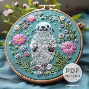 May include: Embroidered hoop art featuring a grey and white seal surrounded by pink and green flowers on a teal background. The text "THE Embroidery Loft" is visible on the hoop.