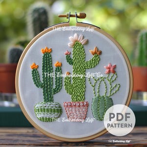 Könnte beinhalten: Stickmuster mit drei Kakteen in Töpfen. Die Kakteen sind grün mit orangefarbenen und rosa Blüten. Das Muster ist in einem Reifen und sagt "THE Embroidery Loft Co" auf dem Stoff.